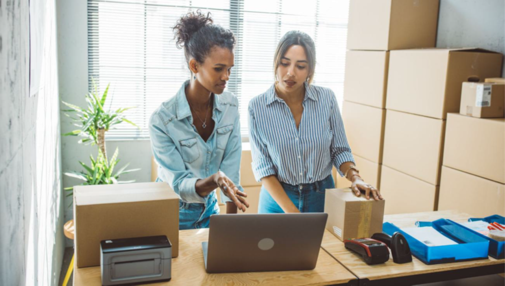 Two women small-business owners work together in a shipping and fulfillment space, reviewing information on a laptop surrounded by boxes, packing supplies, and shipping equipment
